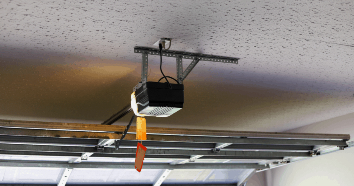 Black, modern garage door opener unit installed on an angle-iron bracket attached to a popcorn ceiling, illuminating the top section of a partially opened sectional garage door.