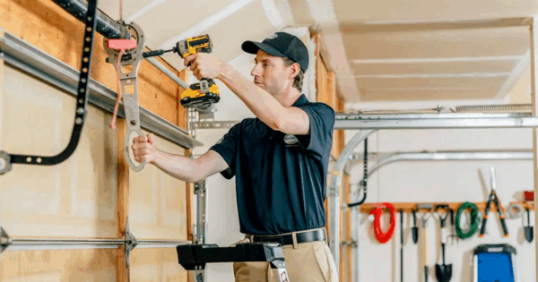 Garage door technician wearing a dark uniform and cap, standing on a stepladder, using a yellow and black power driver to adjust hardware near the torsion spring assembly above a sectional door.